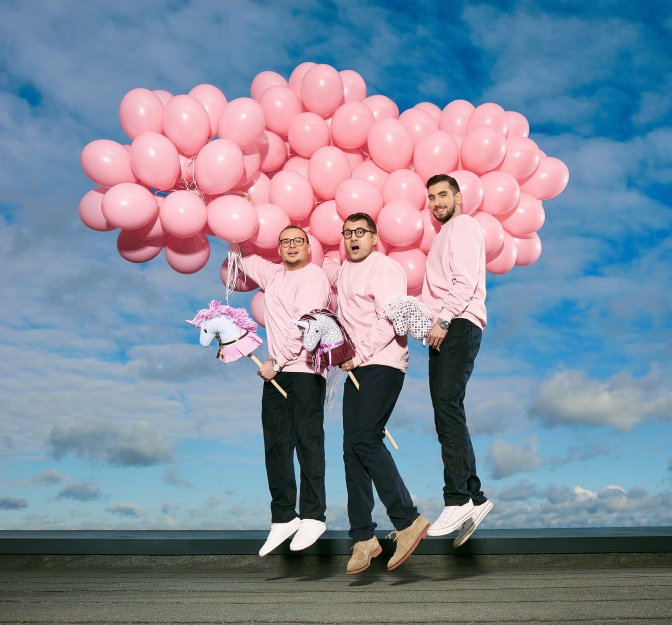 3 Männer in rosafarbenen Sweatshirts halten eine große Traube rosafarbener Luftballons in der Hand.