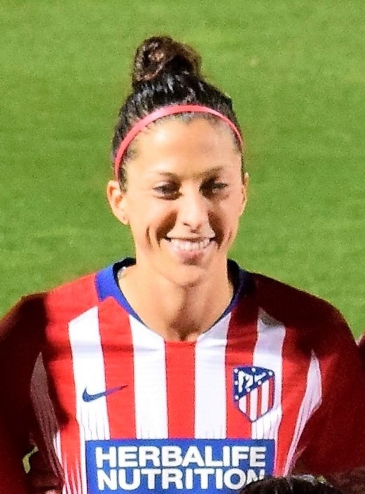 Eine Fußballerin lächelnd auf dem Spielfeld. Sie trägt ein gestreiftes Trikot und hat sich die Haare zu einem Dutt oben auf dem Kopf zusammengebunden.