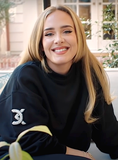 Adele mit langen, glatten blonden Haaren in einem schwarzen Sweatshirt mit Chanel-Logo. Sie lächelt in die Kamera.