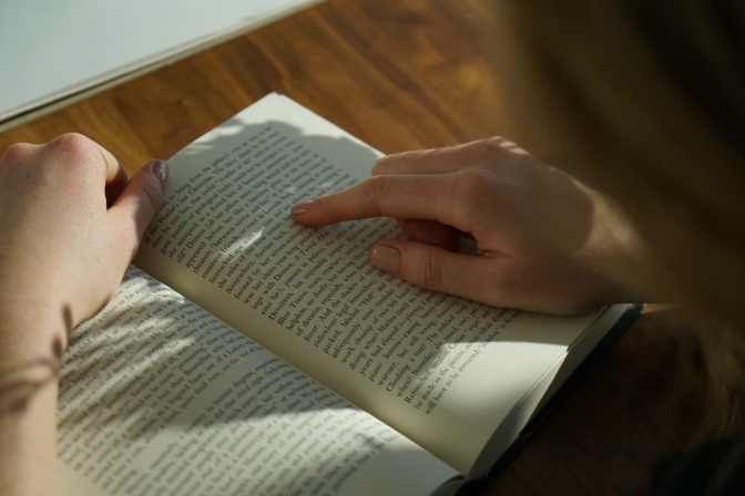 Eine Person liest in einem Buch. Man sieht nur Hände und Hinterkopf. Auf das Buch fallen Schatten einer Pflanze.