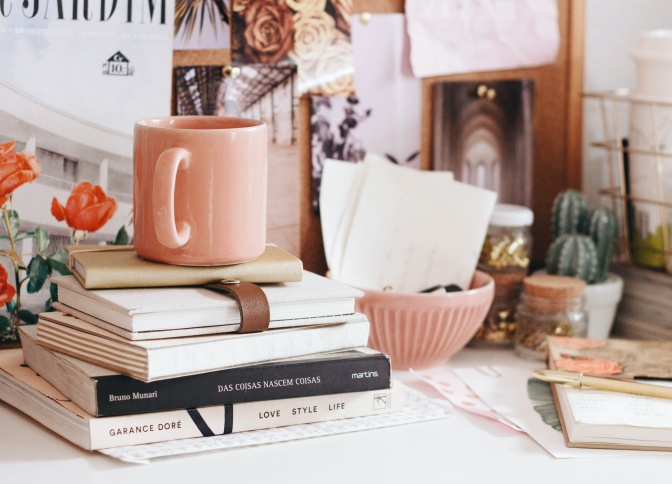 Ein Stapel mit Büchern liegt auf einem Schreibtisch, obenauf steht eine rosane Tasse