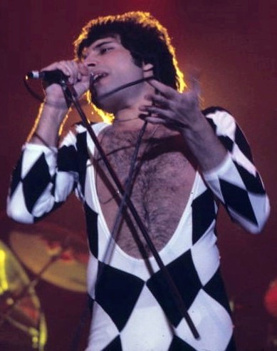 Freddie Mercury in einem Overall mit schwarz-weißem Rautenmuster. Seine Brust ist frei und behaart. Er singt in ein Mikro und gestikuliert mit der Hand.