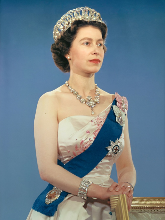 Die Queen im Alter von etwa 30 Jahren in einem festlichen Kleid mit Schärpe, mehreren Orden und Diamantschmuck. Sie schaut ernst seitlich an der Kamera vorbei und hat die Hände übereinandergelegt.