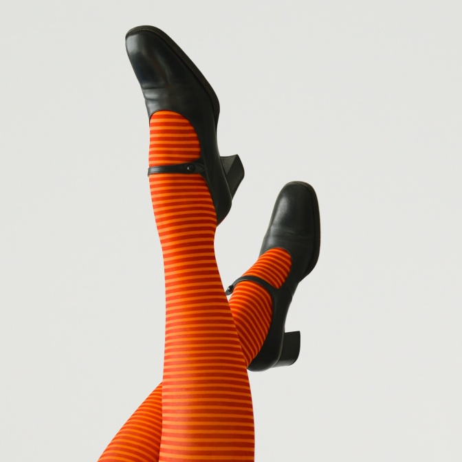 Beine in einer orange-pink geringelten Strumpfhose und schwarzen Riemchenpumps mit runder Spitze