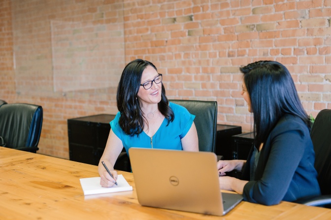 2 Frauen sitzen nebeneinander an einem Tisch. Auf dem Tisch steht ein Laptop. Die beiden haben sich einander zugewendet und sprechen miteinander.