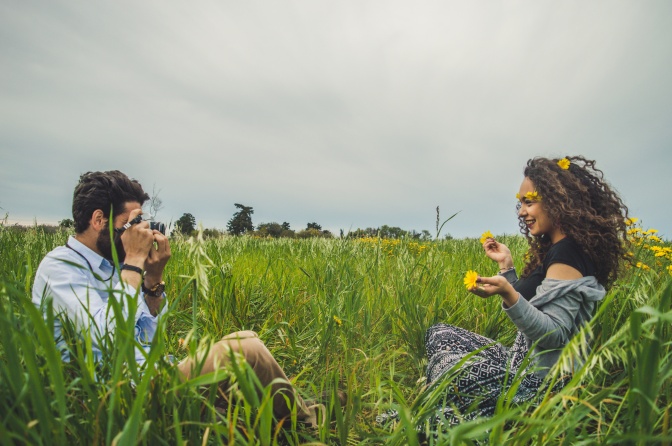 Ein Mann und eine Frau sitzen sich gegenüber auf einer Wiese mit Löwenzahn. Er fotografiert sie mit einer analogen Kamera.