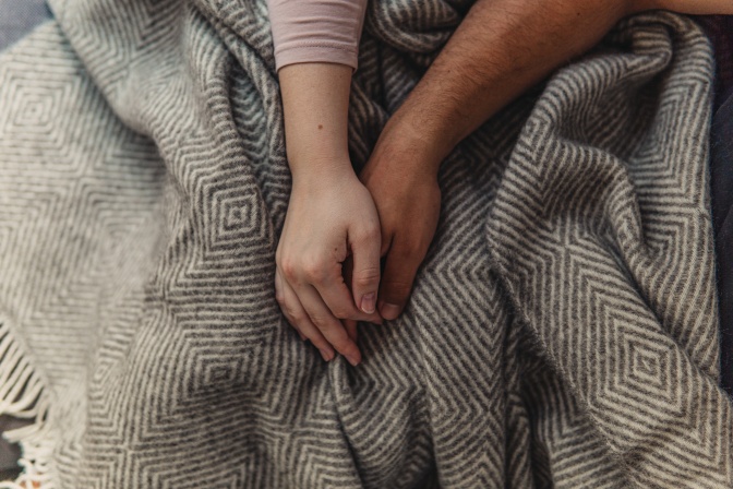 Zwei Menschen halten Händchen. Man sieht nur die Hände und Unterarme auf einer Decke liegen.