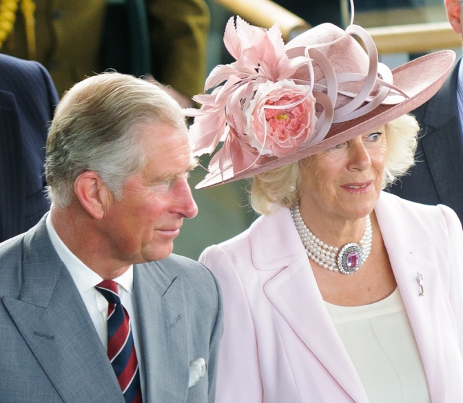 König Charles und Königin Camilla stehen nebeneinander. Sie trägt ein rosa Kostüm mit großem, blumengeschmücktem Hut, er Anzug und eine gestreifte Krawatte.