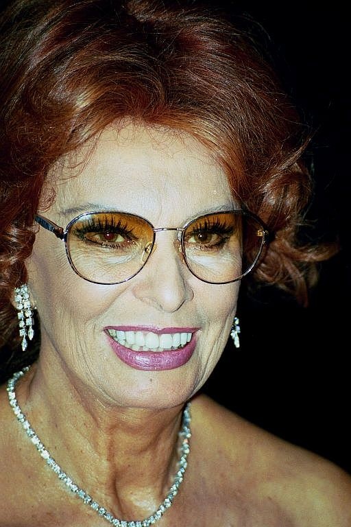 Sophia Loren mit gewellten, kinnlangen roten Haaren. Sie trägt eine große Brille und ist stark geschminkt. Sie lächelt breit.