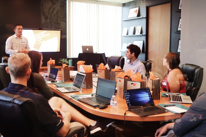 4 Menschen sitzen mit aufgeklappten Laptops um einen Tisch herum. Ein Mann steht vor einem Monitor und hält einen Vortrag.