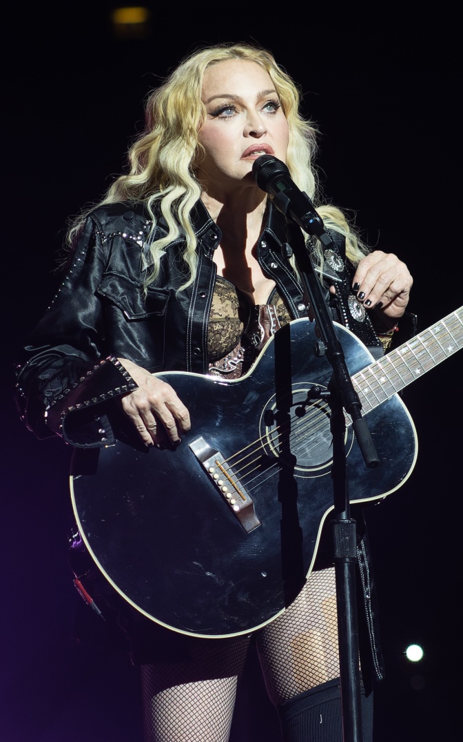 Madonna mit einer schwarzen Akustikgitare auf der Bühne. Sie trägt ein schwarzes Bühnenkostüm und singt in ein Mikrofon.