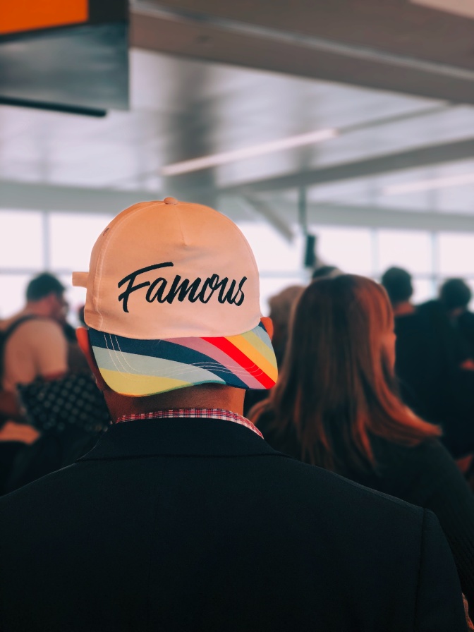 Eine Person mit einer nach hinten gedrehten Baseball-Kappe in Regenbogenfarben. Auf der Kappe steht in geschwungener Schrift das Wort famous, also das englische Wort für berühmt.