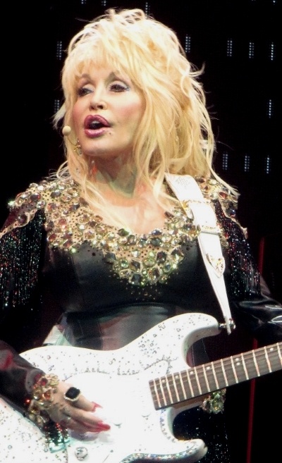Dolly Parton mit einer toupierten und blondierten Frisur auf der Bühne. Sie singt und spielt eine weiße E-Gitarre.