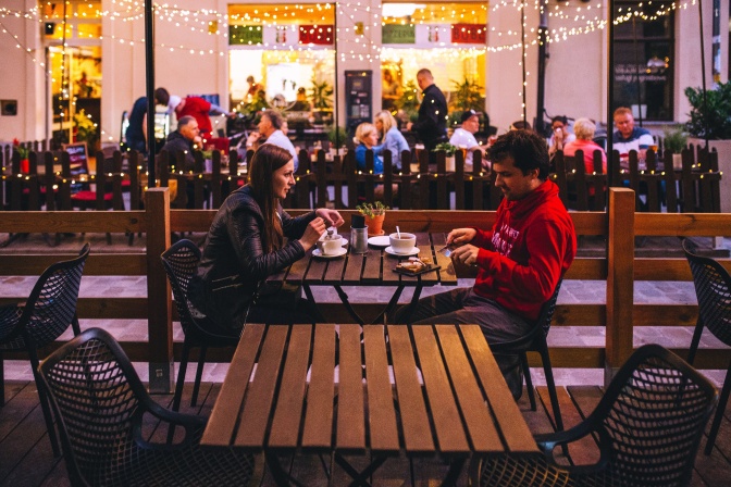 Ein Mann und eine Frau sitzen sich gegenüber an einem Tisch und essen und unterhalten sich. Im Hintergrund viele weitere besetzte Tische im Freien, über Deneen Lichterketten hängen.