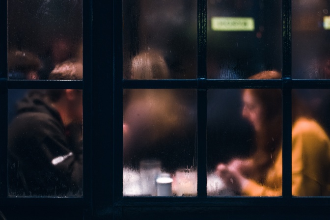 Ein Paar sitzt an einer Kneipe zusammen an einem Tisch. Das Foto ist von außen durch die leicht beschlagene Fensterscheibe fotografiert.