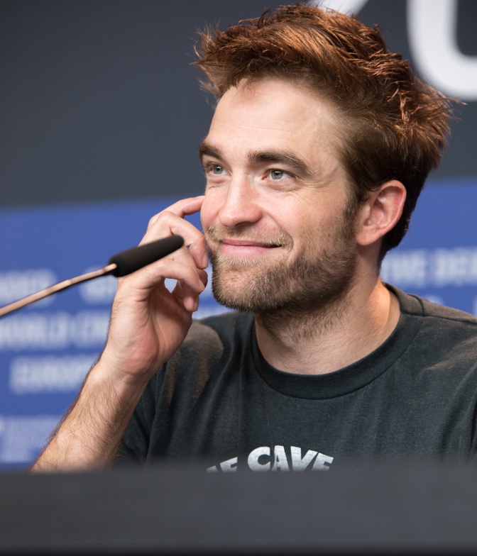 Robert Pattinson sitzt vor einem Mikro und lächelt. Seine braunen Haare stehen ab.