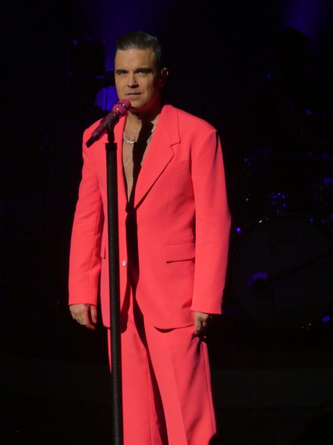 Robbie Williams steht in rotem Anzug vor einem Mikrofon.