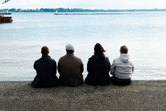 4 Männer mit Down-Syndrom sitzen mit dem Rücken zur Kamera am Meer und schauen aufs Wasser.