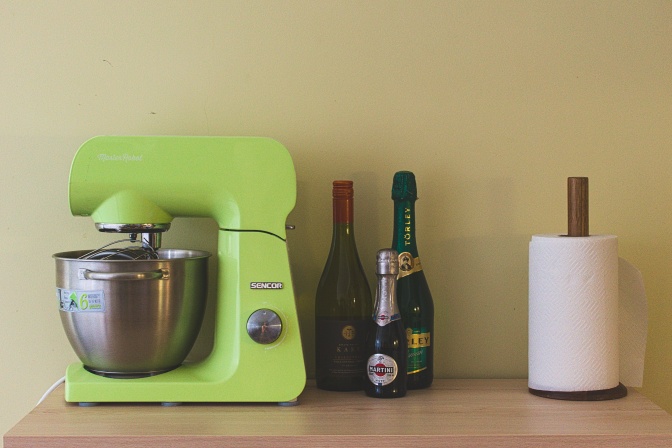 Eine hellgrüne Küchenmaschine steht auf einer Arbeitsfläche neben 2 Flaschen Sekt und einer Küchenrolle.