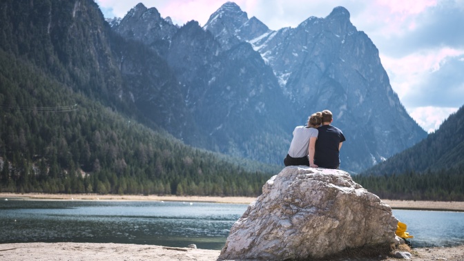 Ein verliebtes Paar sitzt eng beieinander auf einem Stein an einem See. Dahinter sieht man die Berge.