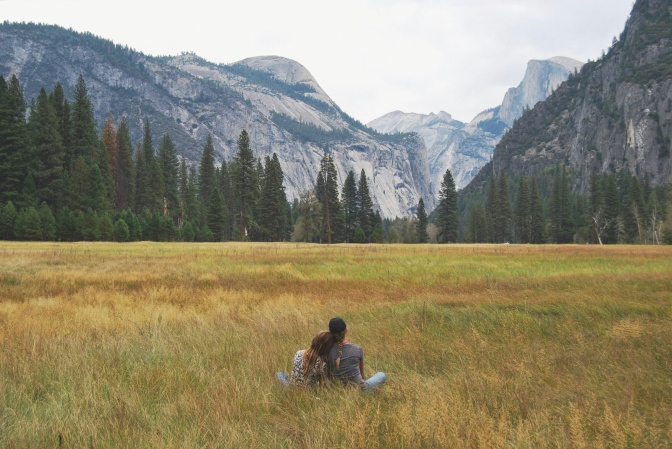 Ein Paar sitzt eng nebeneinander auf einer Wiese, im Hintergrund die Berge.