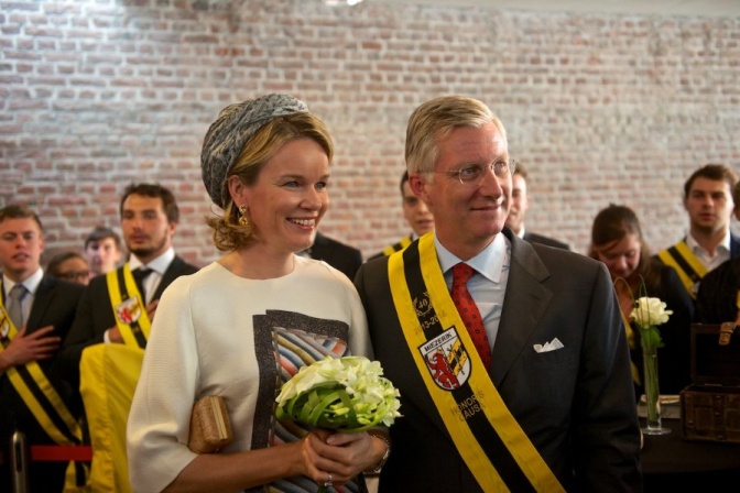 Das Königspaar in festlicher Kleidung vor einer Menschenmenge. Er trägt eine breite Schärpe in den belgischen Landesfarben.
