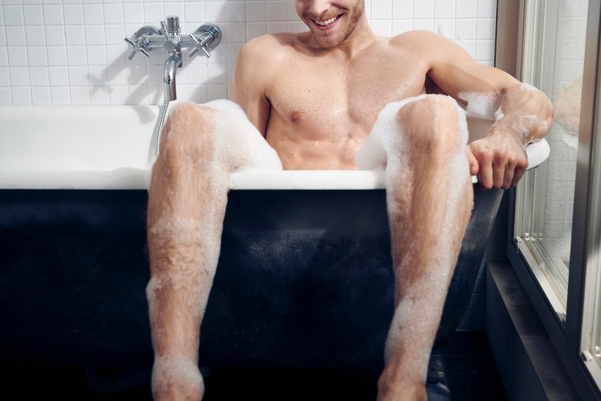 Ein Mann sitzt in einer Badewanne und lässt seine mit Schaum bedeckten Beine aus der Wanne hängen.