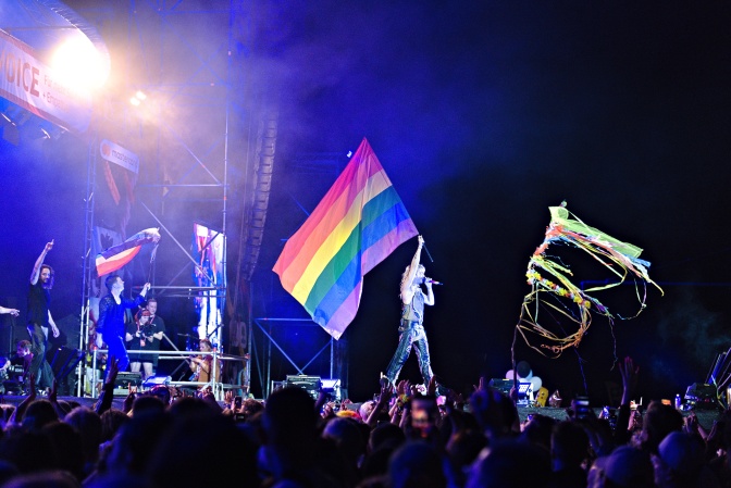 Bandmitglieder auf einer bläulich beleuchteten Bühne. Bill Kaulitz schwenkt eine große Regenbogenflagge.