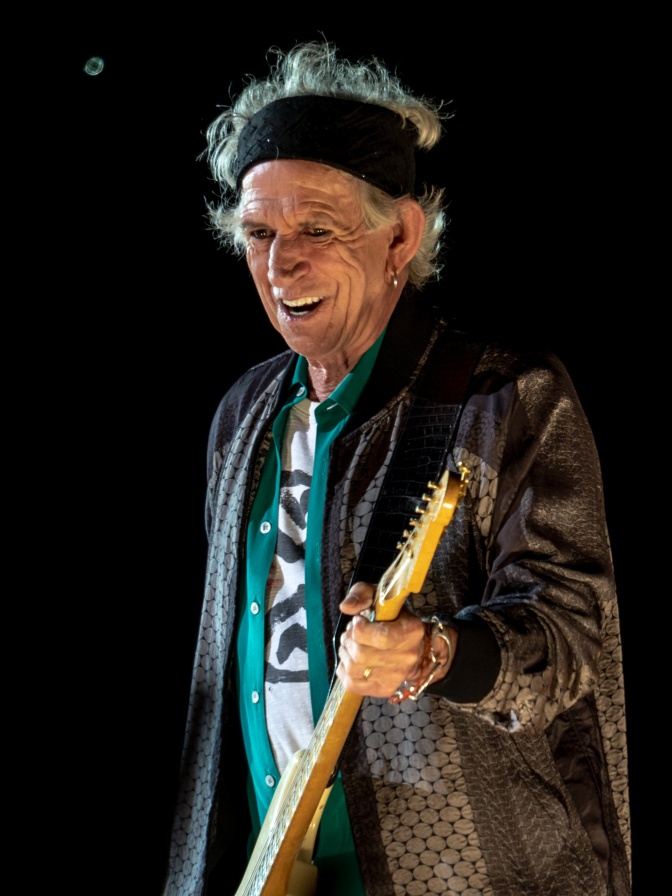 Keith Richards mit Bass auf der Bühne. Er hat graue, gewellte Haare und trägt ein Stirnband.