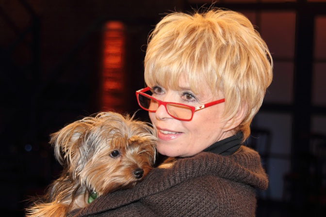 Ingrid Steeger mit kurzen blonden Haaren und einer roten Brille. Sie hält einen kleinen Hund im Arm.
