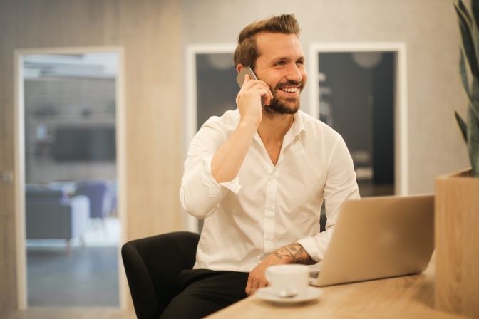 Ein Mann in weißem Hemd sitzt vor einem aufgeklappten Laptop und telefoniert mit dem Handy. Er lächelt dabei.