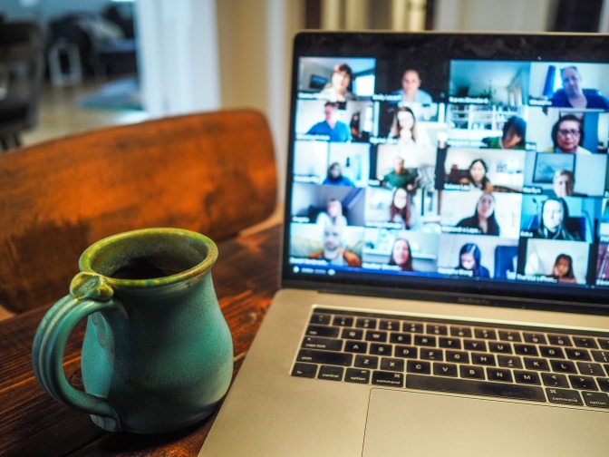 Ein aufgeklappter Laptop zeigt viele Teilnehmer*innen einer Videokonferenz. Neben dem Laptop steht eine Tasse Kaffee.