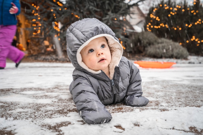 Ein Baby in Schneeanzug mit Kapuze robbt im Freien über den Boden.