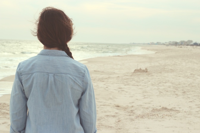 Eine Frau in einem Jeanshemd mit geflochtenem Zopf steht am Meer. Man sieht sie von hinten.