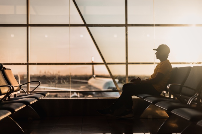 Ein Mann mit Schirmmütze sitzt im Gegenlicht im Wartebereich eines Flughafens. Nur seine Umrisse sind zu sehen, hinter ihm hat der Sonnenuntergang den Himmel orange gefärbt, die Sonne ist aber schon nicht mehr zu sehen,