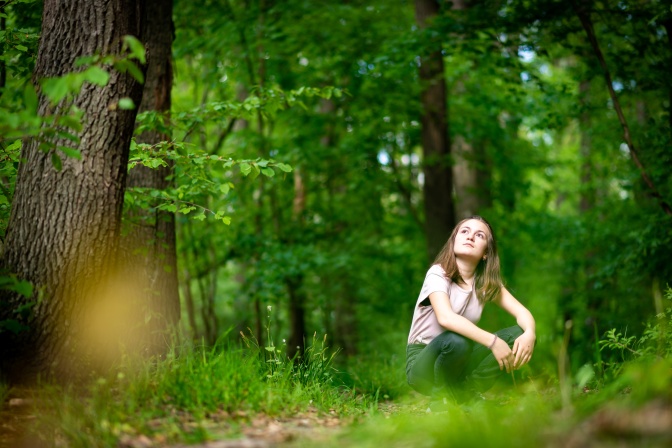 Eine junge Frau kniet im Wald und schaut nach oben ins Grün.