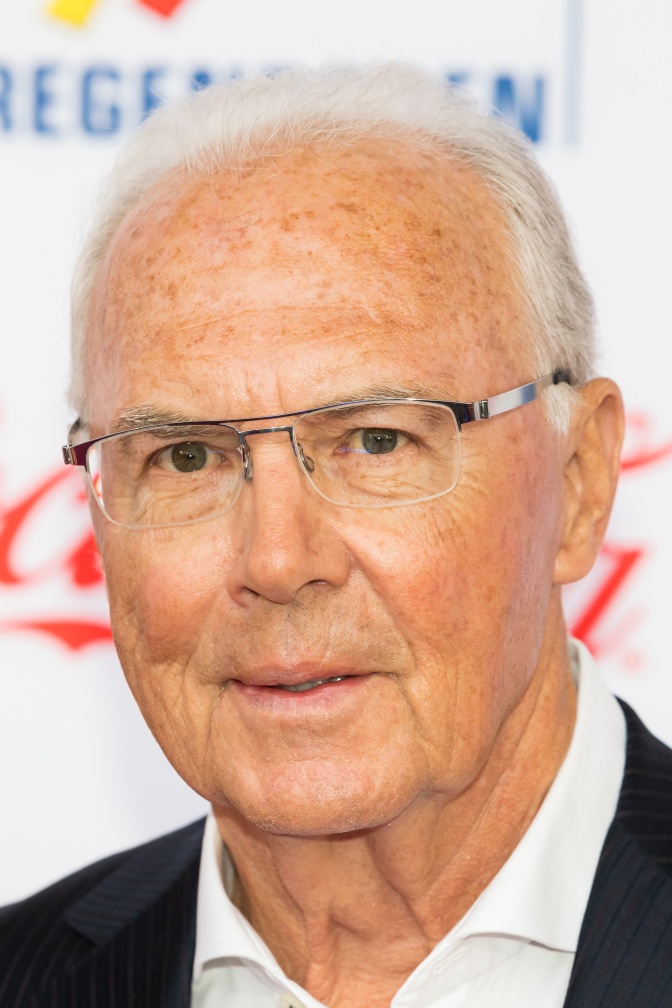 Franz Beckenbauer mit weißen Haaren und randloser Brille vor einer Logowand.