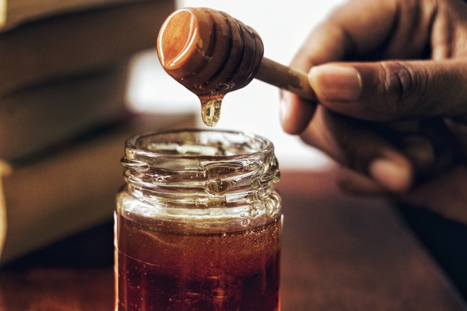 Eine Hand zieht einen Löffel Honig aus einem Glas, der dabei Fäden zieht.