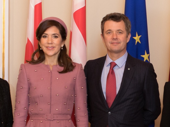 König Frederik von Anzug und Krawatte und seine Frau Königin Mary in einem rosafarbenen Kostüm mit passendem Hut. Hinter den beiden sieht man verschiedene Flaggen.