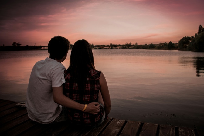 Ein Paar sitzt am Strand. Er legt den Arm um sie. Man sieht die beiden nur im Gegenlicht. Der Sonnenuntergang färbt den Himmel rosa.
