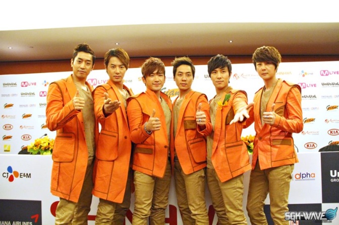 6 koreanische Männer in orangefarbenen Sakkos stehen nebeneinander und schauen in die Kamera.