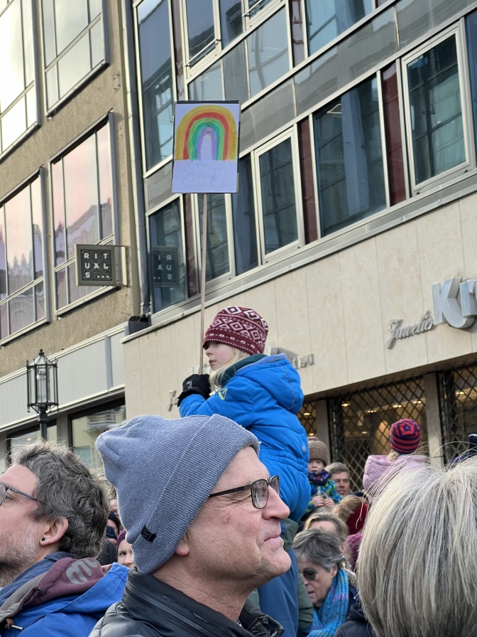 Ein Mann trägt ein Mädchen auf den Schultern. Sie hat ein Demoschild mit einem Regenbogen in der Hand.