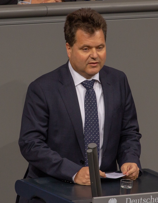 Jürgen Dusel in Anzug und Krawatte. Er hat Textkarten in der Hand und steht vor einem Mikrofon.
