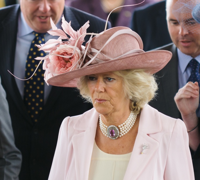 Königin Camilla in einem rosafarbenen Kostüm mit einem sehr großen rosafarbenen Hut mit Blumen.
