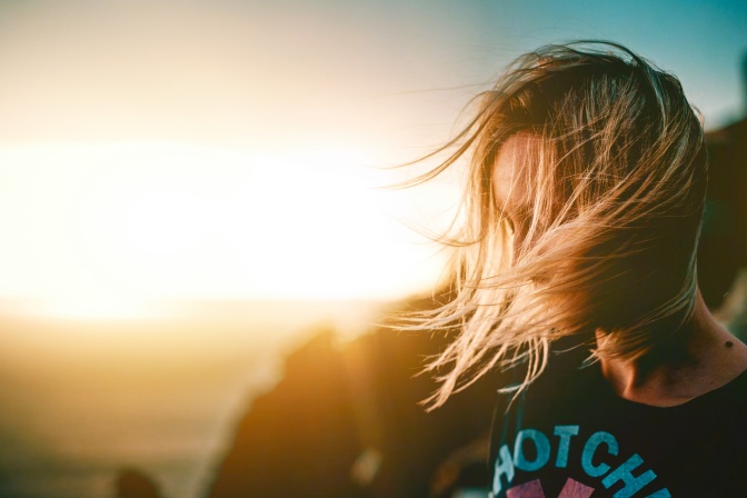 Eine blonde Frau im Gegenlicht des Sonnenuntergangs. Ihr Gesicht wird von ihren im Wind wehenden Haaren verdeckt.