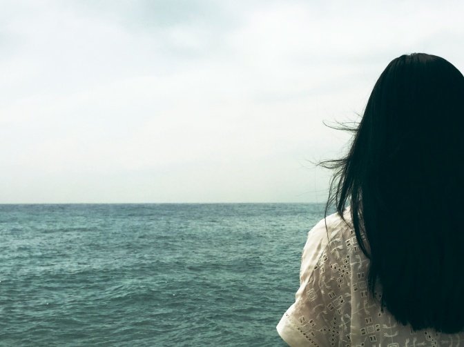 Eine Frau mit schwarzen Haaren am Meer stehend, von hinten fotografiert.