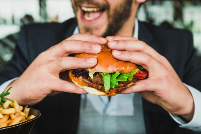 Ein Mann mit Vollbart hält einen Burger in beiden Händen und ist kurz davor, hineinzubeißen.