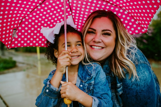 Eine weiße, blonde Mutter mit ihrer schwarzen Tochter unter einem Regenschirm. Das Mädchen ist etwa 4 Jahre alt.