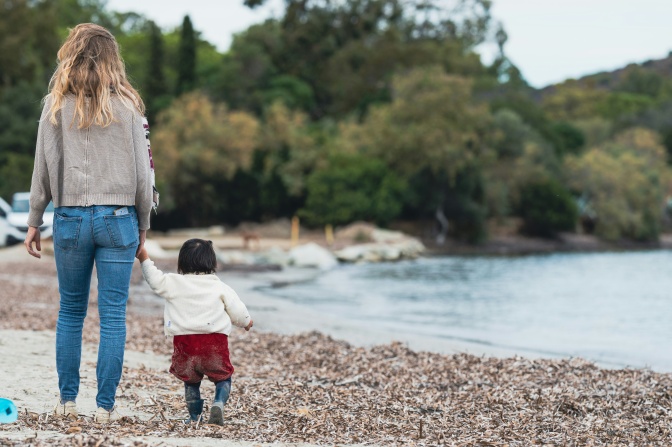 Eine europäisch aussehende Frau hält ein asiatisch aussehendes Kleinkind an der Hand. Beide sind von hinten fotografiert und laufen an einem Ufer entlang.