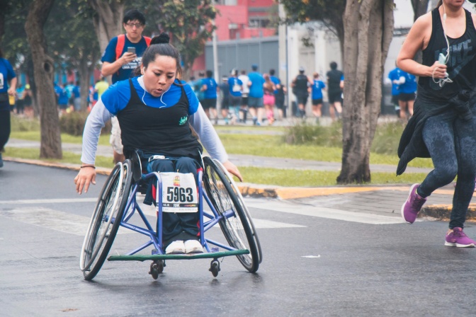 Eine Frau im Rollstuhl nimmt an einem Lauf teil. Sie bewegt den Rollstuhl mit angestrengtem Gesicht vorwärts. Am Rollstuhl ist eine Startnummer befestigt.